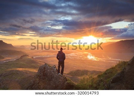 Man standing on a ledge of a mountain, enjoying the beautiful sunset over a wide river valley in Thorsmork, Iceland.