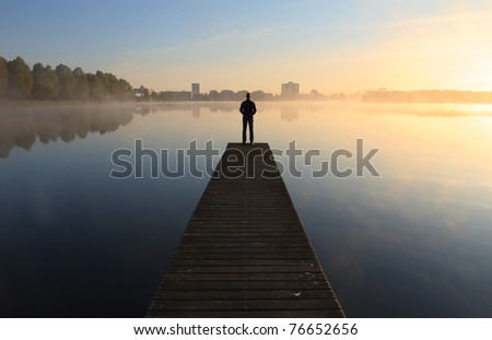 Man on a pier looking toward the city over a foggy lake during sunrise.