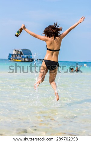 Sexy Asian Thai girl in bikini is jumping with joy on the seashore of Pattaya sea with alcohol bottle on her hand and water splashing for summer vacation concept
