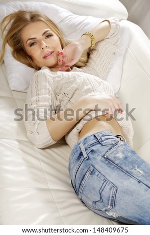 Beautiful and attractive young adult smile blonde woman posing in blue jeans and grey sweater on the white sofa