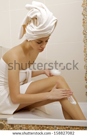 Attractive young adult woman applying moisturizer cream on the legs in bathroom