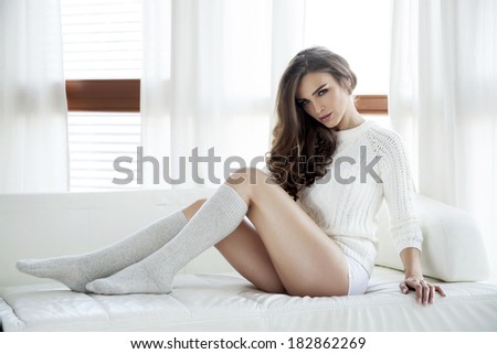 Beautiful and sexy young adult sensuality brunette woman in white sweater and grey socks on the white sofa in window on luxury apartment