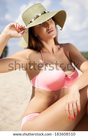 Young adult beautiful brunette woman in pink bikini and green hat on the beach