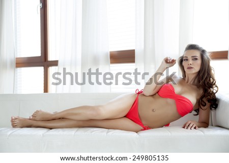 Beautiful and sexy young adult pretty sensuality brunette woman in red bikini on the white sofa and sunny window in luxury style apartment