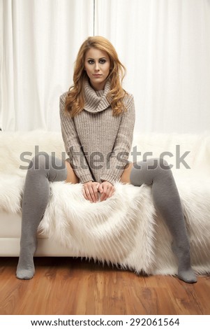 Beautiful and sexy young adult sensuality blonde woman in gray sweater and white socks on the white sofa in fur