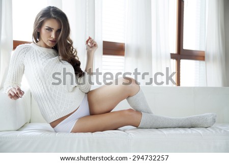 Beautiful attractive and sexy young adult sensuality pretty brunette woman in white sweater and grey socks on the white couch in sunlight window on luxury apartment