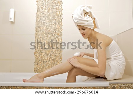 Portrait of a beautiful attractive young adult pretty brunette woman applying moisturizer cream on the legs in bathroom