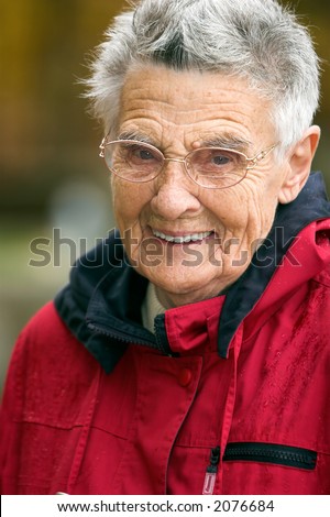 Smiling senior woman in red jacket with short hair wearing glasses against a blurry background. Narrow DOF with focus on the eyes.