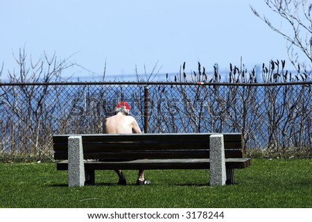 A shirtless man sitting alone on a park bench overlooking a lake.