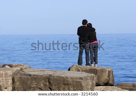 Loving couple hugging, while standing on a rock looking out over the water on a sunny afternoon.