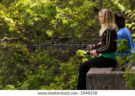 Pretty blond woman fishing while sitting on a concrete ledge on the bank of a river.