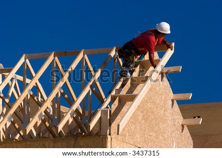 Carpenter with red shirt and white hardhat building a roof of a house at a construction site.