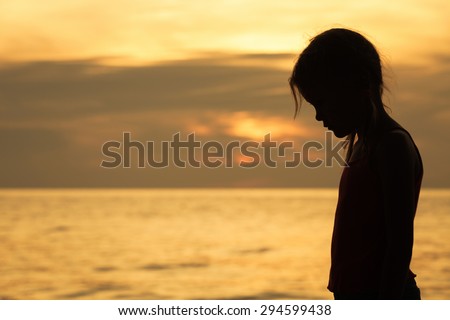Portrait of sad blond little girl standing on the beach at sunset time.