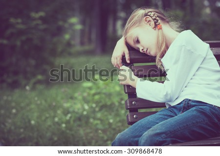 Portrait of sad blond little girl sitting on bench at the day time.