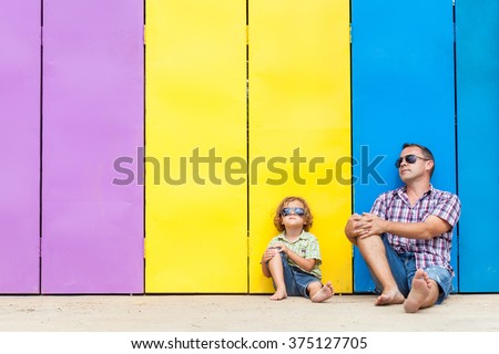 Father and son relaxing near the house at the day time. They sitting near are the colorful wall. Concept of friendly family.