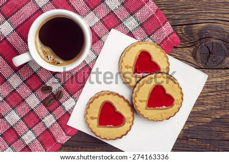 Cookies with jam in shape of hearts and cup of black coffee on red tablecloth, top view