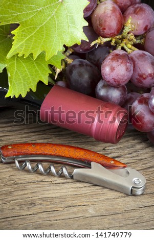 A corkscrew is in front of wine bottle and grapes with vine leaves on rustic wooden background