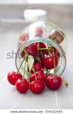 Freshly picked ripe cherries with stem and leaves in front of a preserving jar filled with cherries