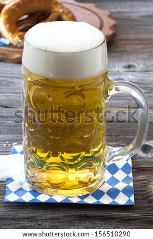 Large beer mug with beer on a napkin with blue and white rhombuses on a rustic wooden table in the background a wooden cutting board with pretzels