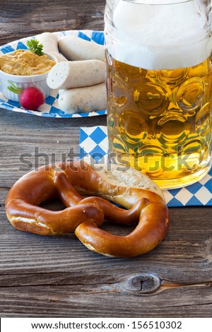 A cold beer in Seidel, a pretzel in front and in the background a paper plate with veal sausages and mustard