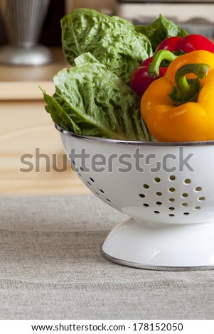 Fresh vegetables in a white enamel colander in the background a kitchen cupboard