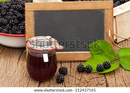 Blank slate blackboard in front of ripe blackberries and a jar blackberry jelly on a rustic wooden table