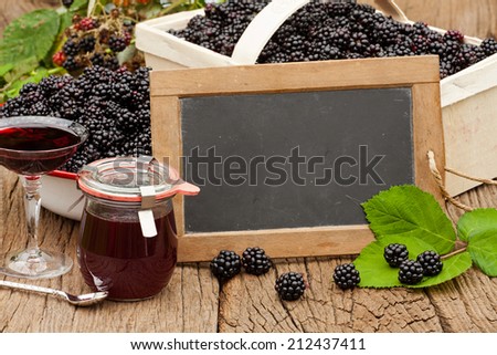 Blank slate blackboard in front of ripe blackberries, blackberry liqueur and a jar blackberry jelly on a rustic wooden table