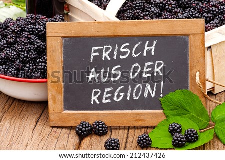 Slate blackboard with the Germans words: Frisch aus der Region (Fresh from the region), in front of ripe blackberries on a rustic wooden table
