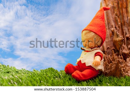 Seated doll dressed elf sitting on green grass against the trunk of a tree on a sunny day