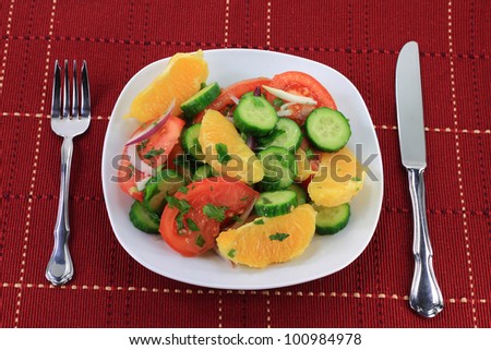 Salad: Vegetables Ã¢Â?Â? cucumbers, tomatoes; Fruit Ã¢Â?Â? orange; Seasoning - red onion, cilantro; Dressing: lime juice, olive oil in white porcelain dish and silverware Ã¢Â?Â? fork, knife over red serviette.