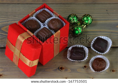 Chocolate candies in cups into red box and lid decorated with gold ribbon as well spilled out over wooden table and three green Balls for Christmas Decoration.