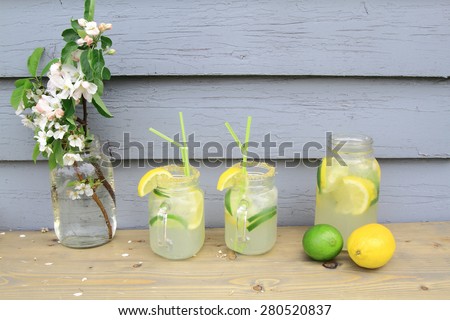 In hot summer on the table decorated with apple tree blooms in backyard is served cold Lemonade in decorated with crystals brown sugar jar glasses, Ice cubes, slices lemon limes and drinking straws