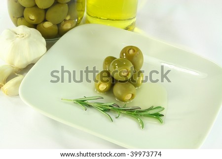Still life picture represent green stuffed with garlic pickled olives in jar and served on dish with olive oil and rosemary.