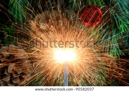 Picture of burning (lit) sparkler focal point and green Christmas tree, pine cones and decorative red ball as blurred background.