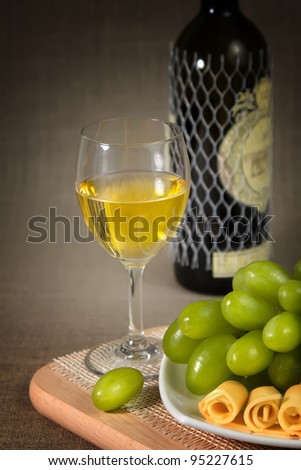 A glass of white wine, green grapes, cheese and a bottle on a wooden surface