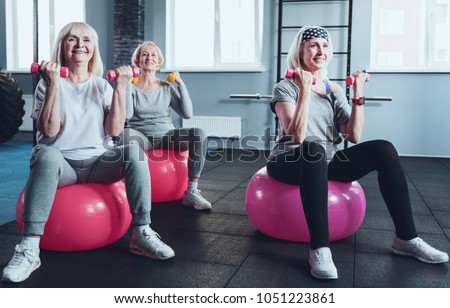Say no to a passive lifestyle. Group of three radiant ladies focusing on an exercise while all sitting on fitness balls and lifting dumbbells during a training session.