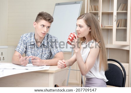 Young fair-haired architect talking to his beautiful colleague wearing nice white blouse and drinking coffee. White board for copy place behind them