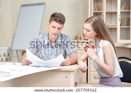 Two young architects sitting at the table with different plans and schemes on it discussing their new project. White board for copy place behind them