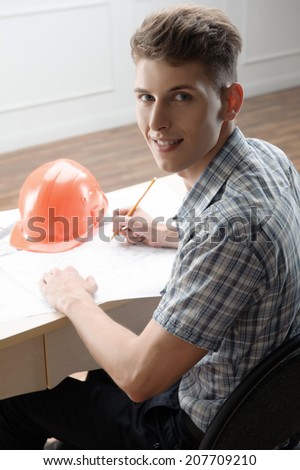 Young fair-haired smiling architect sitting at the table with different plans and schemes and orange helmet on it sketching his new project and looking at us