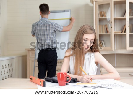 Selected focus on beautiful fair-haired architect working at the new project. Her young colleague wearing checked shirt and black pants measuring the length of white board for copy place on background
