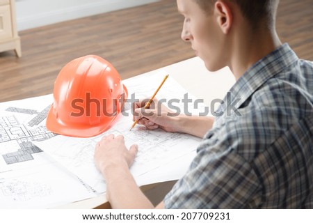 Young fair-haired architect sitting at the table with different plans and schemes and orange helmet on it sketching his new project