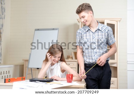 Young architect wearing checked shirt standing with the rule near the table laughing with his nice colleague. White board for copy place behind them
