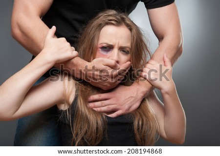 Frightened young girl with the black eye wearing black shirt wanted to get free from her heavy-handed husband but she cannot. Isolated on grey background
