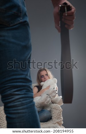 Man standing back to us and holding girdle in his hands looking at the young scared girl with black eye sitting on the chair and keeping big teddy bear. Isolated on grey background