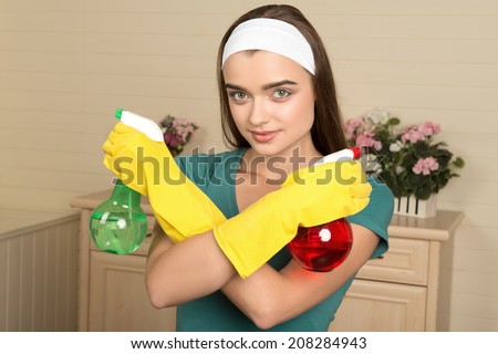 Half-length portrait of beautiful dark-haired housemaid wearing white fillet with crossed arms holding two colorful water sprayers and looking at us