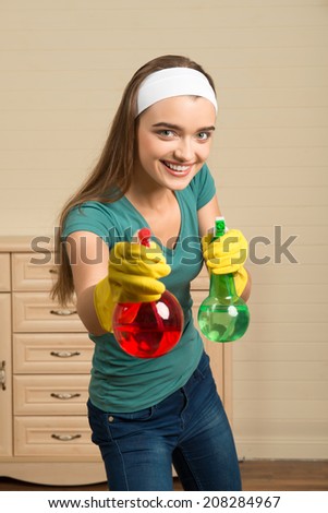 Half-length portrait of pretty young smiling housemaid wearing white fillet and yellow rubber gloves holding two colorful water sprayers. She is ready to clean