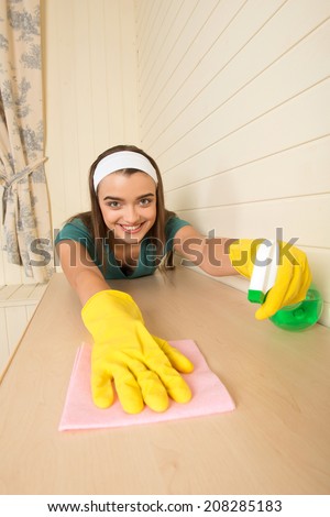 Lovely dark-haired housemaid with the white fillet wearing yellow gloves wiping cream-colored table in the kitchen