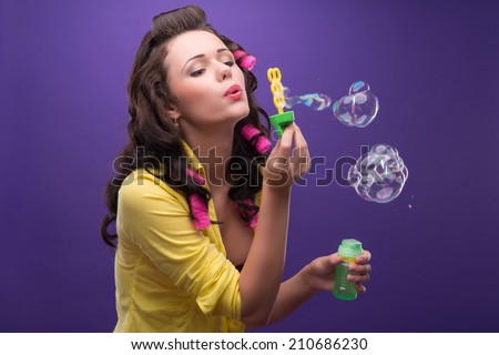 Half-length portrait of charming young smiling woman with curly hair wearing nice yellow shirt and pink curlers blowing the bubbles. Isolated on blue background