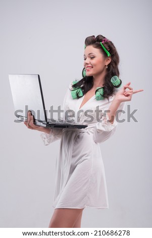 Half-length portrait of beautiful young smiling woman wearing green curlers and white satin dressing gown standing aside to us with the notebook looking at something very funny. Isolated on blue