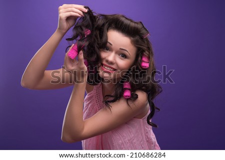 Half-length portrait of charming young smiling woman with curly hair wearing nice pink dress and cannot take off the curlers. Isolated on blue background